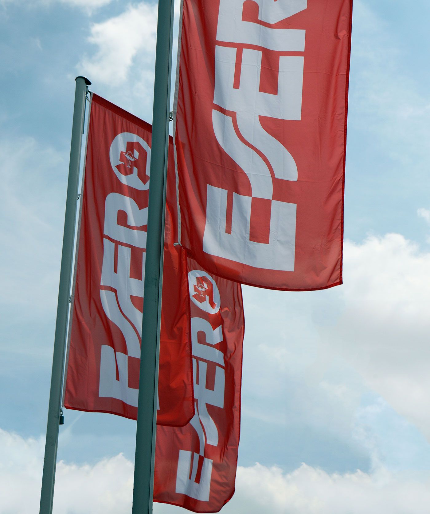 Fotografie mehrerer roter Werbefahnen an Metallmasten vor blauem Himmel mit Wolken. Weiße, teilweise gedrehte Schriftzüge und ein rundes Emblem prägen die Fahnen; Wind lässt sie unterschiedlich weit auswehen.